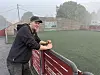 Ein mann støttar seg på eit raudt gjerde ved ein kunstgrasbane med murvegg og gråver i bakgrunnen.